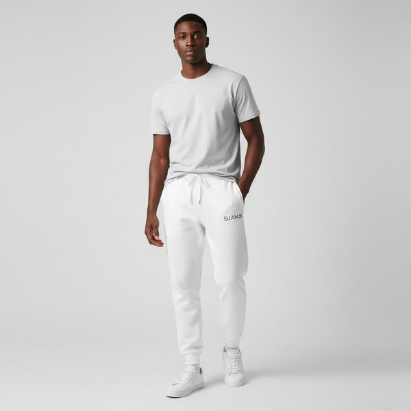 Man standing in white B.I.AM.B sweatpants with a small logo on the left thigh, paired with a light gray T-shirt and white sneakers on a plain studio background.