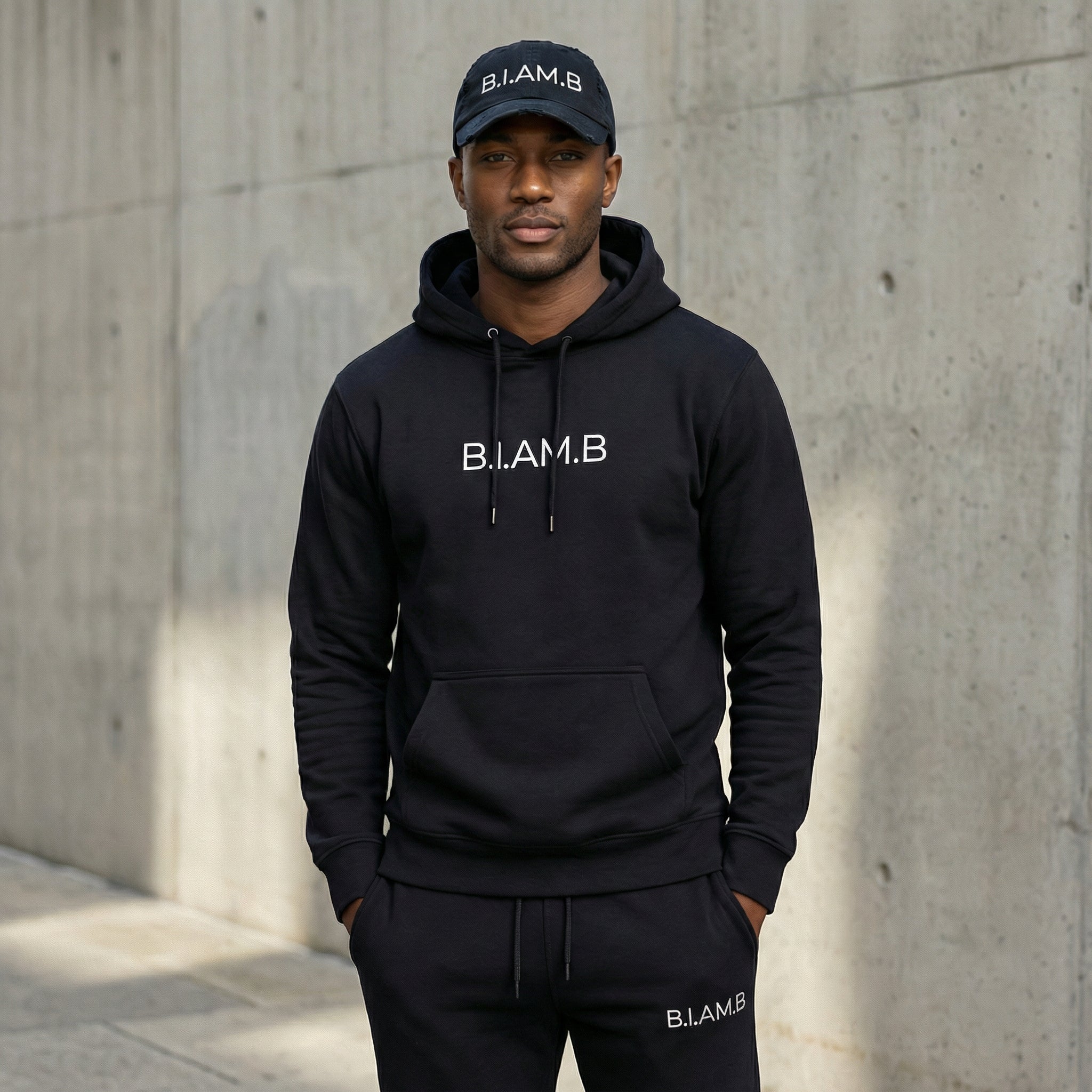 Man wearing B.I.AM.B black hoodie and black cap with logo, lifestyle streetwear look against concrete wall